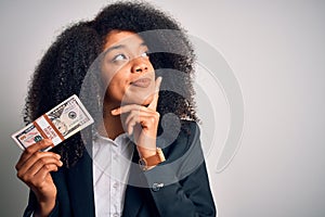 Young african american business woman with afro hair holding a bunch of cash dollars banknotes serious face thinking about