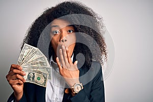 Young african american business woman with afro hair holding cash dollars banknotes cover mouth with hand shocked with shame for