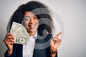 Young african american business woman with afro hair holding cash dollars banknotes very happy pointing with hand and finger to