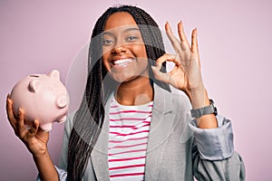 Young african american business woman saving money on piggy bank over isolated background doing ok sign with fingers, excellent