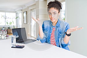Young african american business woman working using computer laptop clueless and confused expression with arms and hands raised