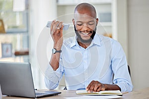 Young african american businessman on a call using a phone while reading a note in a notebook and working on a laptop in