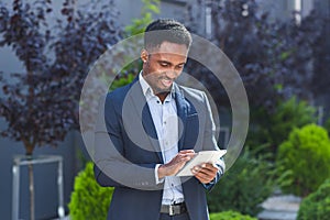 Young african american businessman in formal business suit standing working with tablet in hands on background modern office