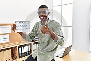 Young african american businessman holding travel calendar at the office smiling happy and positive, thumb up doing excellent and