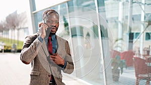 Young African American businessman stands next to the modern office talks on his smartphone. He talks to his business