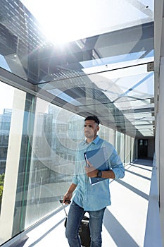 Young african american businessman walking with file and luggage at airport corridor on sunny day