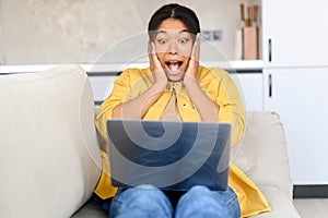 Young African American businesswoman with Afro hairstyle, eyes wide open covering mouth with hands, sitting at the sofa