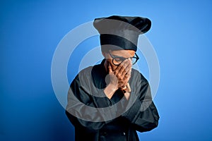 Young african american chef woman wearing cooker uniform and hat over blue background with sad expression covering face with hands