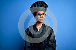 Young african american chef woman wearing cooker uniform and hat over blue background smiling looking to the side and staring away