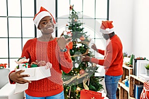Young african american couple standing by christmas tree smiling with happy face looking and pointing to the side with thumb up