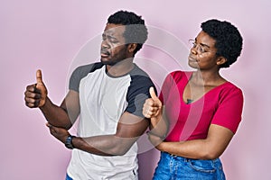 Young african american couple standing over pink background looking proud, smiling doing thumbs up gesture to the side
