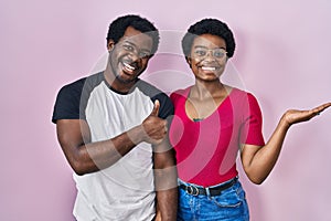 Young african american couple standing over pink background showing palm hand and doing ok gesture with thumbs up, smiling happy