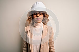 Young african american curly architect woman wearing safety helmet and glasses with serious expression on face