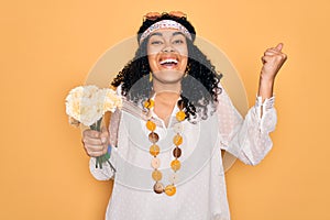 Young african american curly hippie woman wearing sunglasses holding bouquet of flowers screaming proud and celebrating victory