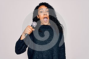 Young african american curly singer woman singing using microphone over white background scared in shock with a surprise face,