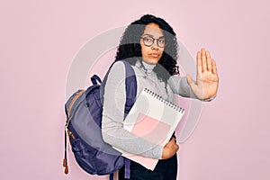 Young african american curly student woman wearing backpack and glasses holding book with open hand doing stop sign with serious