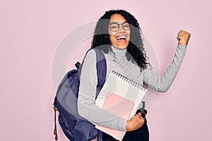 Young african american curly student woman wearing backpack and glasses holding book screaming proud and celebrating victory and