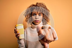 Young african american curly woman drinking cup of takeaway coffee over yellow background pointing with finger to the camera and