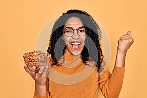 Young african american curly woman holding bowl with healthy almonds over yellow background screaming proud and celebrating