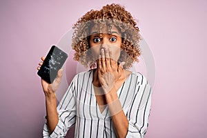 Young african american curly woman holding broken smartphone showing craked screen cover mouth with hand shocked with shame for