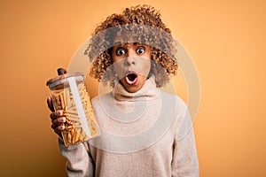 Young african american curly woman holding jar with Italian dry pasta macaroni scared in shock with a surprise face, afraid and