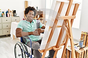 Young african american disabled artist man holding paintbrushes sitting on wheelchair at art studio