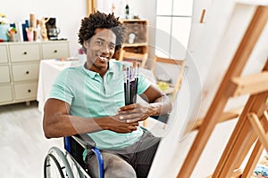 Young african american disabled artist man holding paintbrushes sitting on wheelchair at art studio