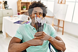 Young african american disabled artist man holding paintbrushes sitting on wheelchair at art studio