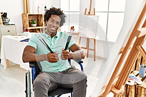 Young african american disabled artist man holding paintbrushes sitting on wheelchair at art studio