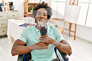 Young african american disabled artist man holding paintbrushes sitting on wheelchair at art studio