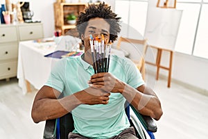 Young african american disabled artist man holding paintbrushes sitting on wheelchair at art studio