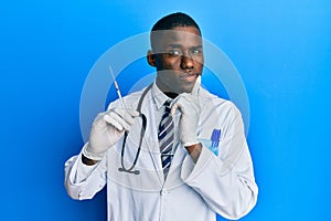 Young african american doctor man holding syringe serious face thinking about question with hand on chin, thoughtful about