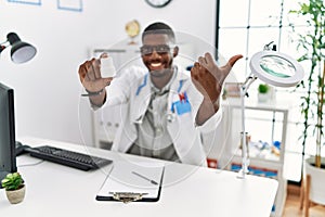 Young african american doctor man wearing doctor uniform holding prescription pills at the clinic pointing thumb up to the side