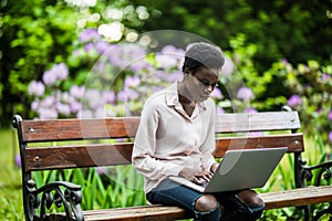 Young African-American female with the laptop is sitting on the wooden bench. cheerful cute black girl on the park bench using the