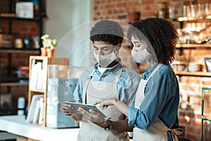 Young african american female and male in aprons and protective masks check financial accounts with digital tablet