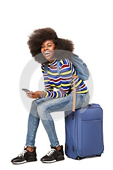 Young african american female traveler sitting on suitcase with mobile phone