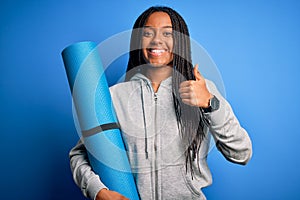 Young african american fitness woman holding yoga mat over  blue background happy with big smile doing ok sign, thumb up