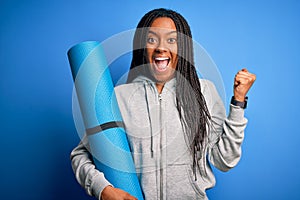 Young african american fitness woman holding yoga mat over isolated blue background screaming proud and celebrating victory and