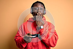 Young african american gamer man playing video game using joystick and headphones cover mouth with hand shocked with shame for