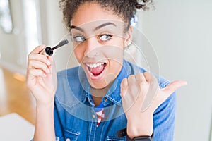 Young african american girl applying eyelashes mascara pointing and showing with thumb up to the side with happy face smiling