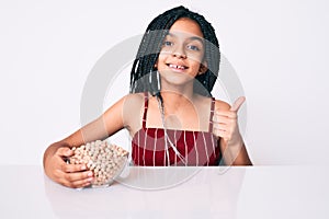 Young african american girl child with braids holding chickpeas bowl smiling happy and positive, thumb up doing excellent and