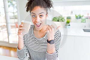 Young african american girl drinking a coffee on a take away paper cup pointing and showing with thumb up to the side with happy