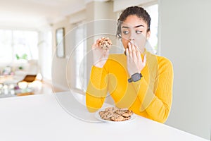 Young african american girl eating chocolate chips cookies as sweet snack cover mouth with hand shocked with shame for mistake,