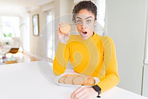 Young african american girl eating healthy whole grain biscuits scared in shock with a surprise face, afraid and excited with fear