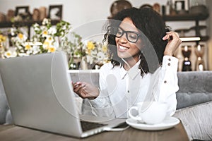 Young African American girl in glasses sitting in restaurant with laptop. Pretty girl with earphones joyfully looking in