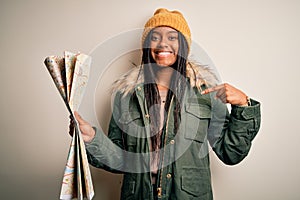 Young african american girl going on vacation holding turist city map over  background with surprise face pointing finger