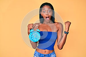 Young african american girl holding alarm clock screaming proud, celebrating victory and success very excited with raised arms