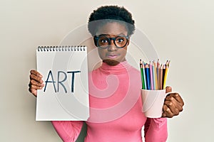 Young african american girl holding art notebook and colored pencils puffing cheeks with funny face