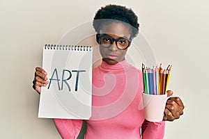 Young african american girl holding art notebook and colored pencils relaxed with serious expression on face