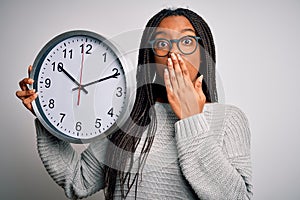 Young african american girl holding big minute clock over isolated background cover mouth with hand shocked with shame for